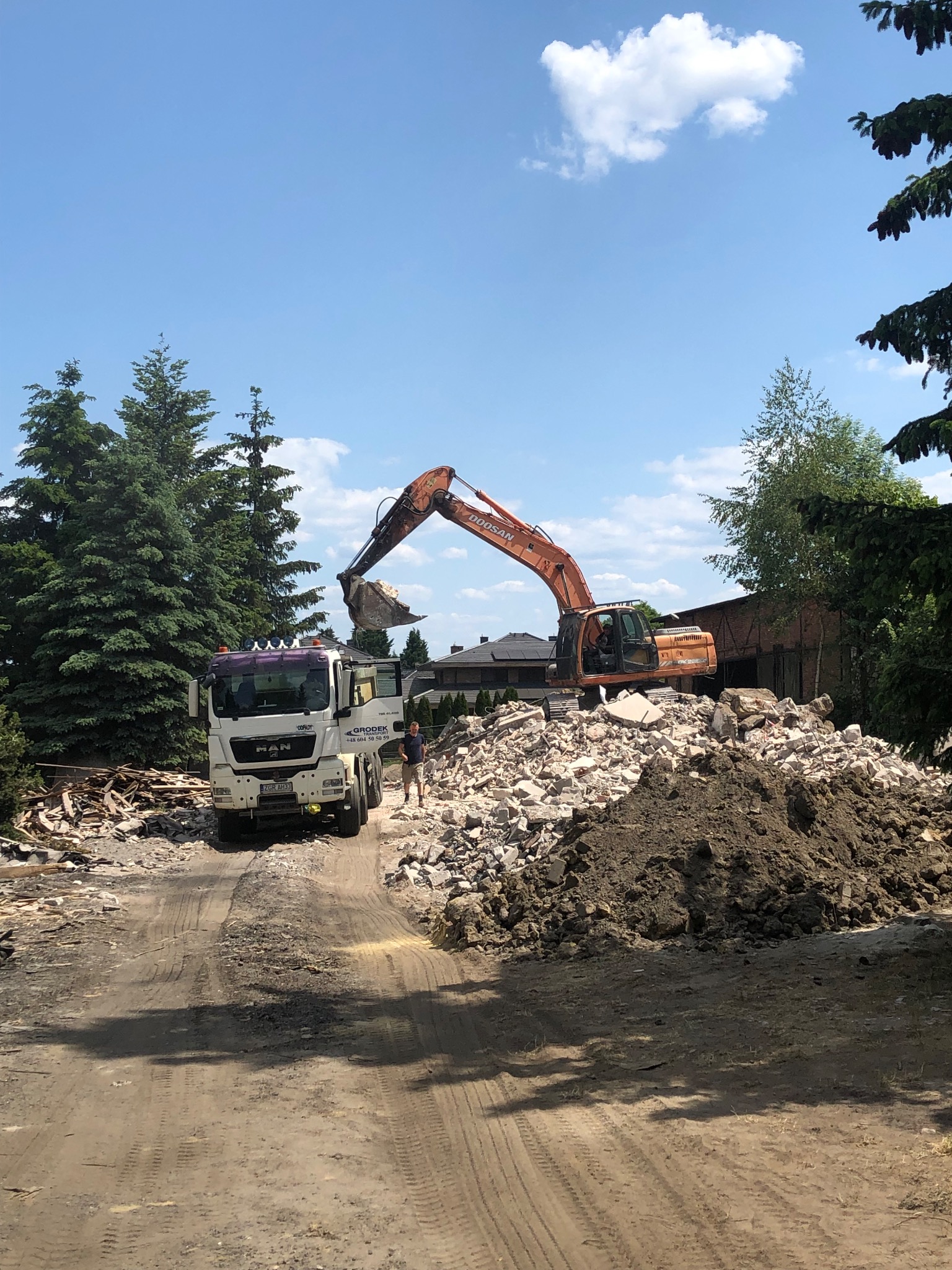 Pomarańczowa koparka Doosan ładuje gruz na ciężarówkę MAN z otwartymi drzwiami, na tle drzew i błękitnego nieba z chmurami.