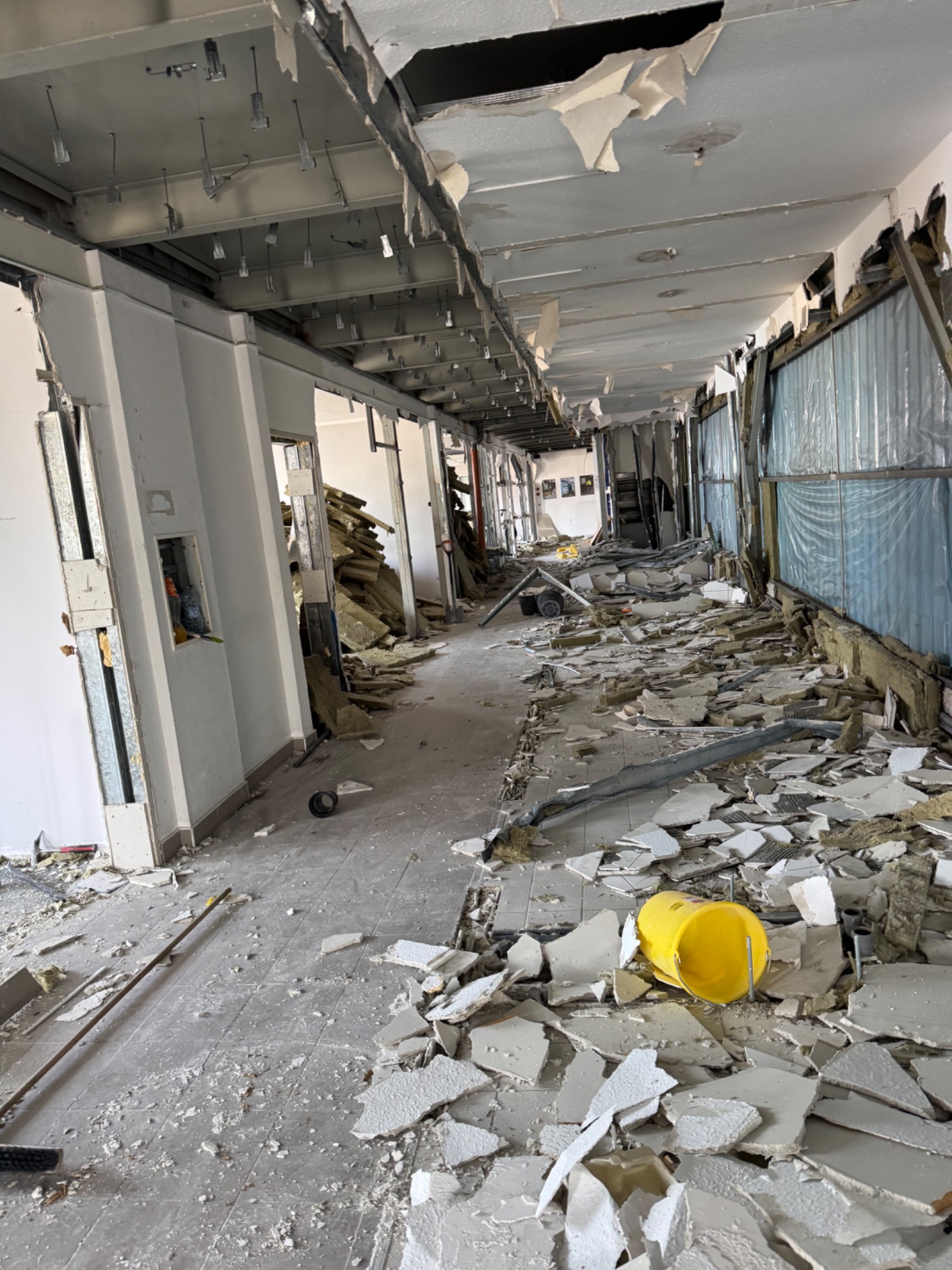 Wnętrze po demontażu ścian i sufitu podwieszanego, gruz na podłodze, widoczne elementy konstrukcyjne budynku i instalacje. Żółte wiadro na pierwszym planie.