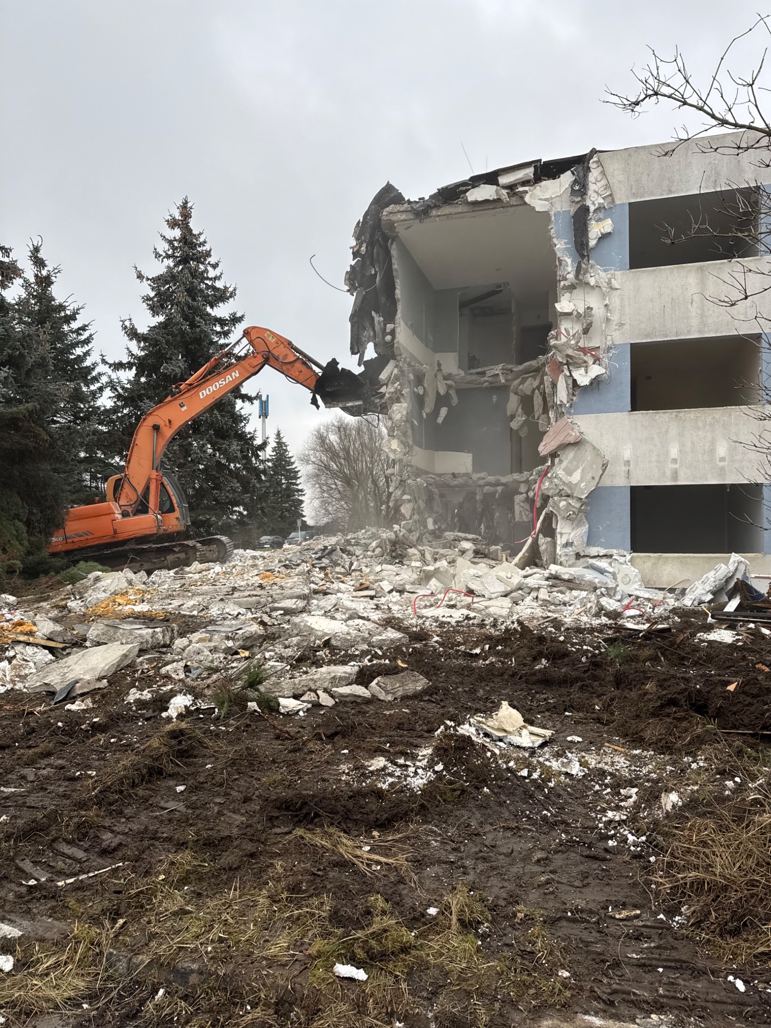 Pomarańczowa koparka Doosan w trakcie rozbiórki częściowo zburzonego, wielopiętrowego budynku z elementami niebieskiej elewacji, otoczonego gruzem i zielenią.