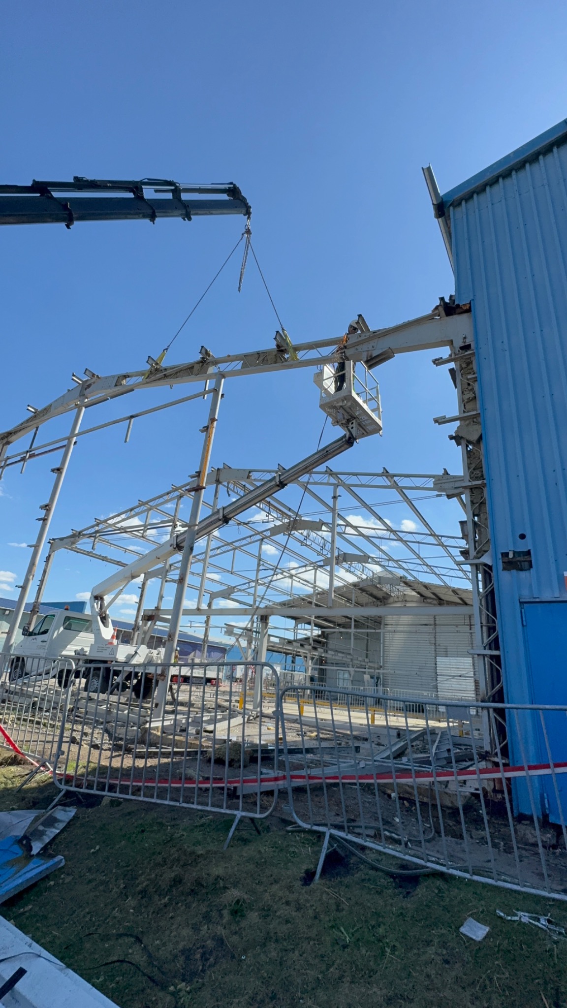 Demontaż konstrukcji stalowej hali przez dźwig i podnośnik koszowy. Widoczne elementy konstrukcji, pracownicy i fragment niebieskiej ściany budynku.