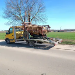 Żółta laweta przewozi stary opryskiwacz rolniczy w Turzynie. Na tle pola i pojedynczego wiatraka.
