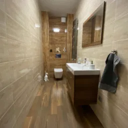 Wąska łazienka z beżowymi płytkami imitującymi drewno na ścianach i podłodze, wisząca szafka z umywalką, toaleta pod ścianą, grzejnik drabinkowy i dekoracje w kształcie kaczek.