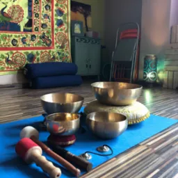 Cztery misy dźwiękowe z pałeczkami na niebieskiej macie w studio z drewnianą podłogą, widoczne poduszki do siedzenia i dekoracyjna tkanina z motywem pawia w tle.