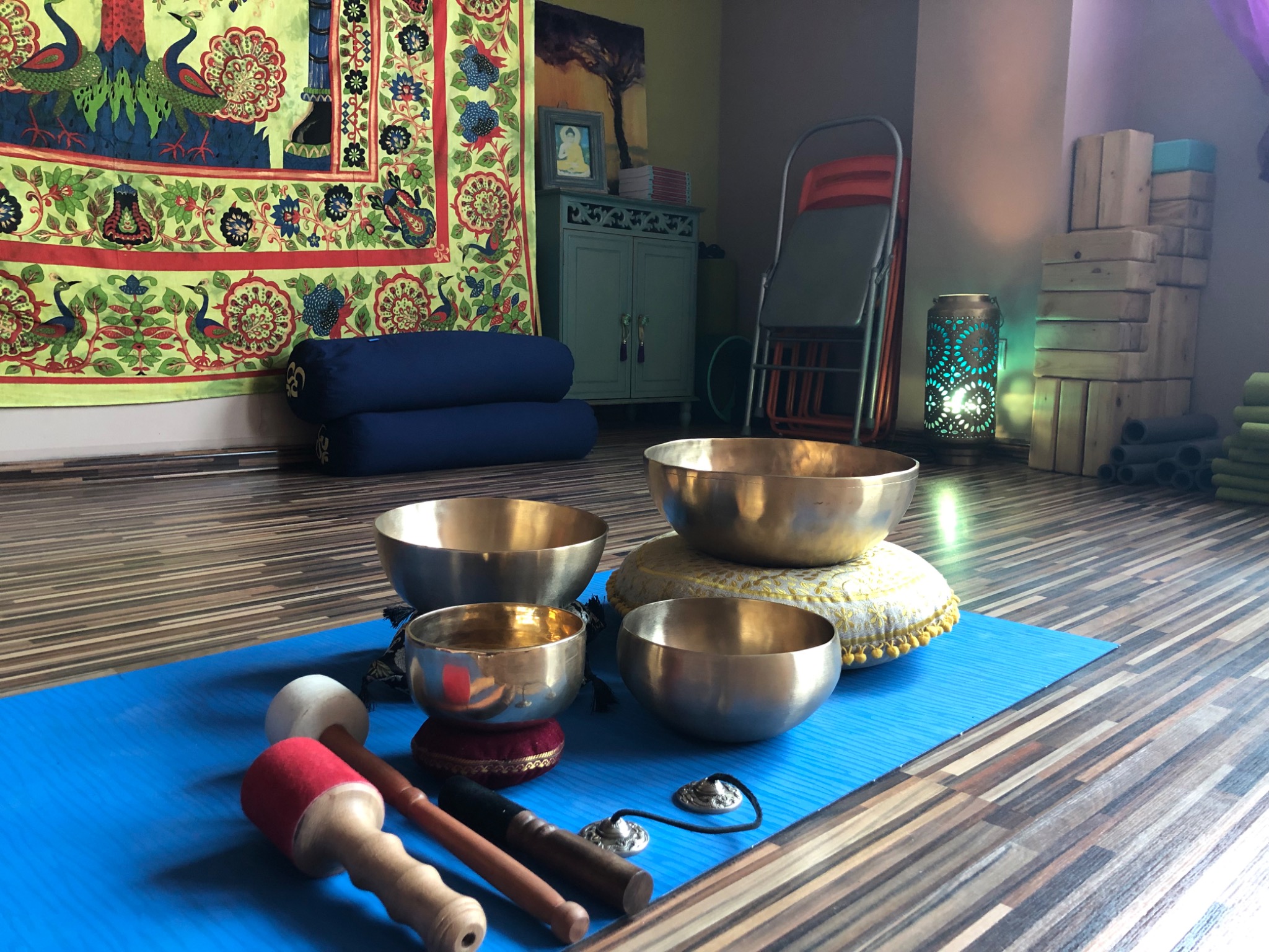 Cztery misy dźwiękowe z pałeczkami na niebieskiej macie w studio z drewnianą podłogą, widoczne poduszki do siedzenia i dekoracyjna tkanina z motywem pawia w tle.