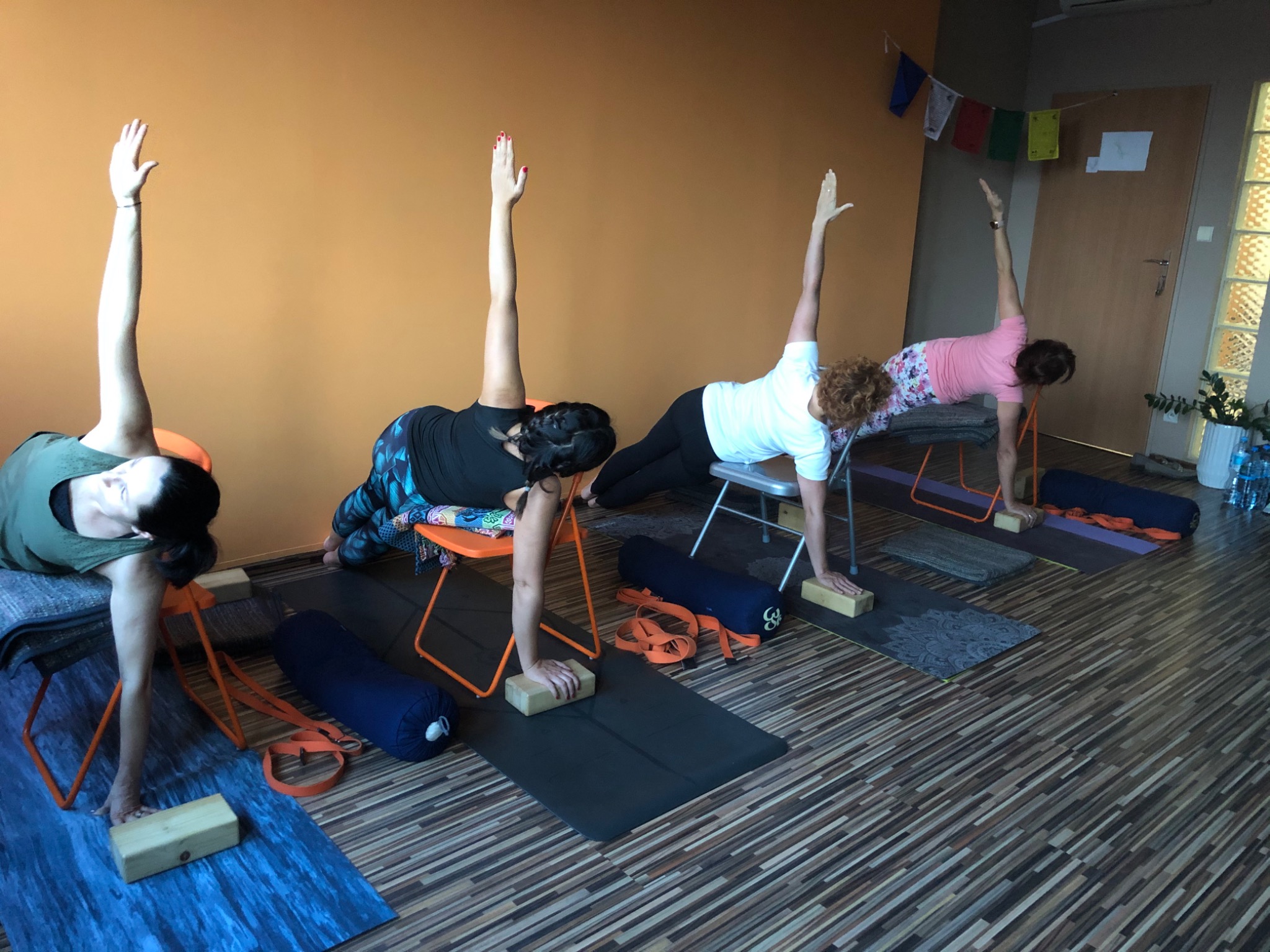 Grupa osób ćwiczy pozycję boczną na krzesłach w studiu, używając drewnianych klocków i mat do ćwiczeń.