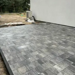 Ułożona, szara kostka brukowa z czarnym obrzeżem przy ścianie budynku z klimatyzatorem. Częściowo przygotowany teren pod dalsze prace ziemne.