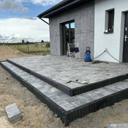 Mateusz Piotrowski - Ułożony taras z kostki brukowej w odcieniach szarości, z dwoma stopniami obrzeżowanymi czarnym krawężnikiem, na tle nowoczesnego domu i błękitnego nieba.