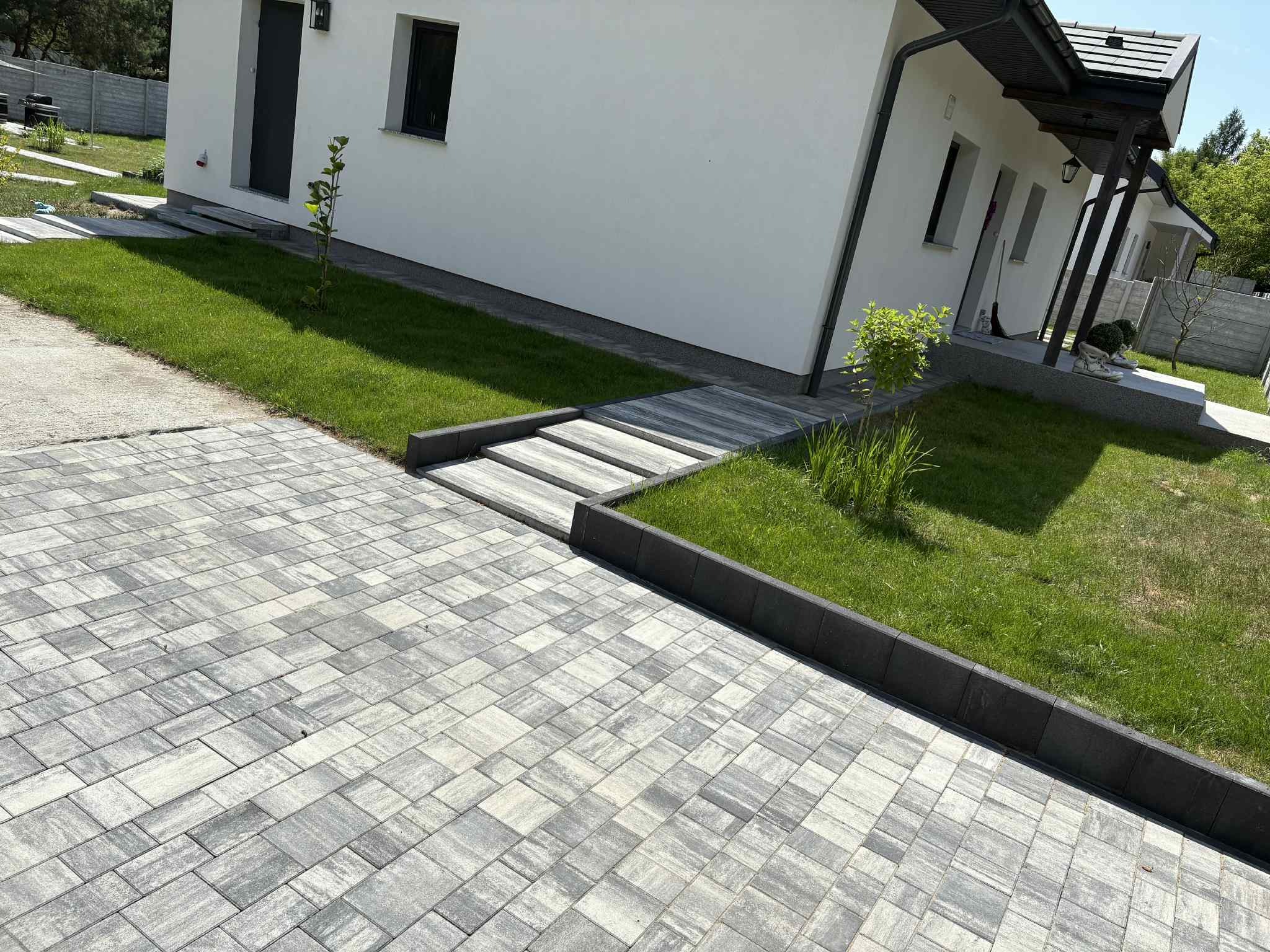 Nowoczesny podjazd z szarej kostki brukowej prowadzący do białego domu z zadbanym trawnikiem i elementami małej architektury ogrodowej, widok z perspektywy pieszego.