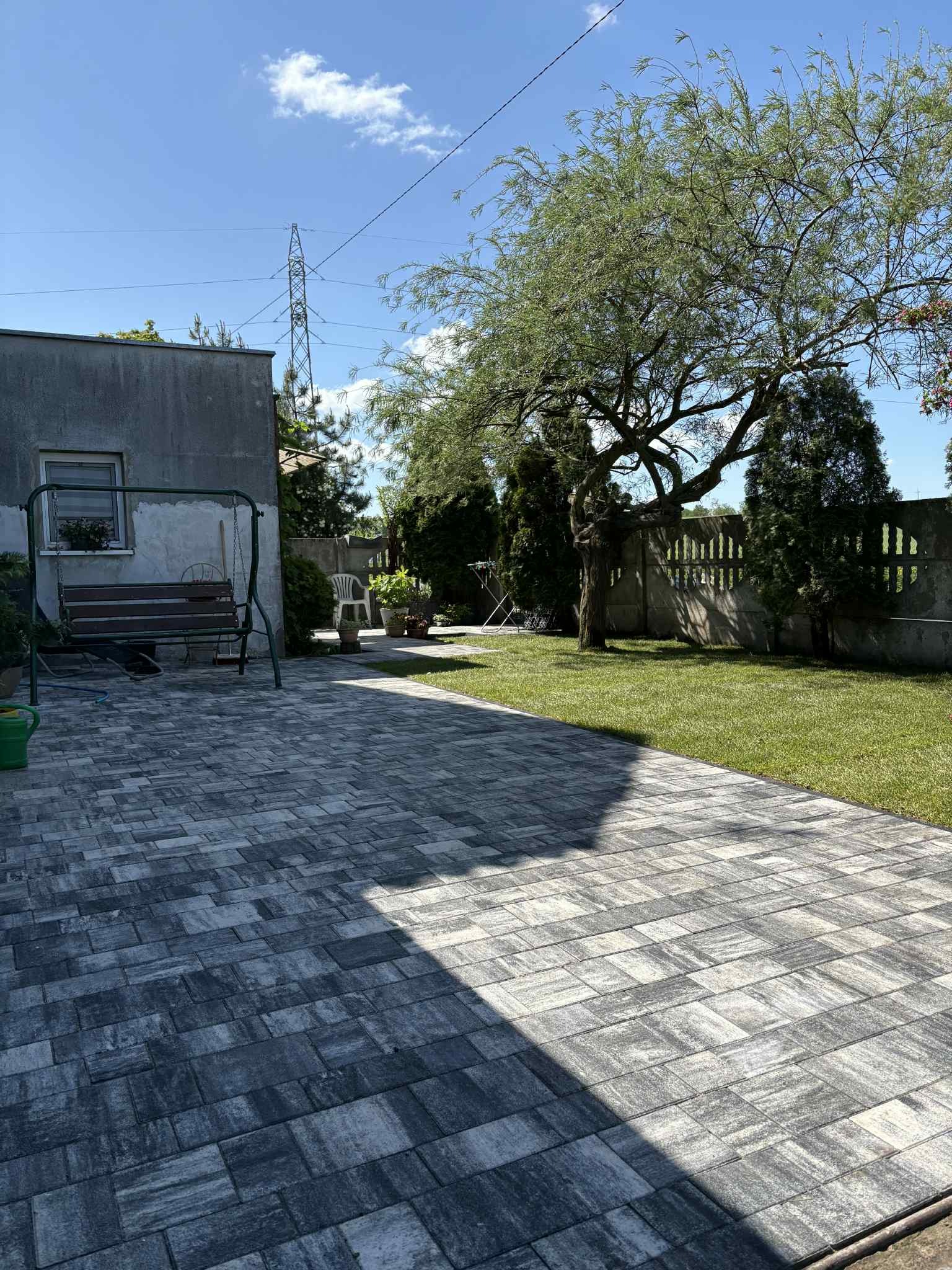 Ułożona kostka brukowa w odcieniach szarości tworzy ścieżkę w ogrodzie. W tle dom, trawnik, drzewa i betonowe ogrodzenie. Na pierwszym planie cień.