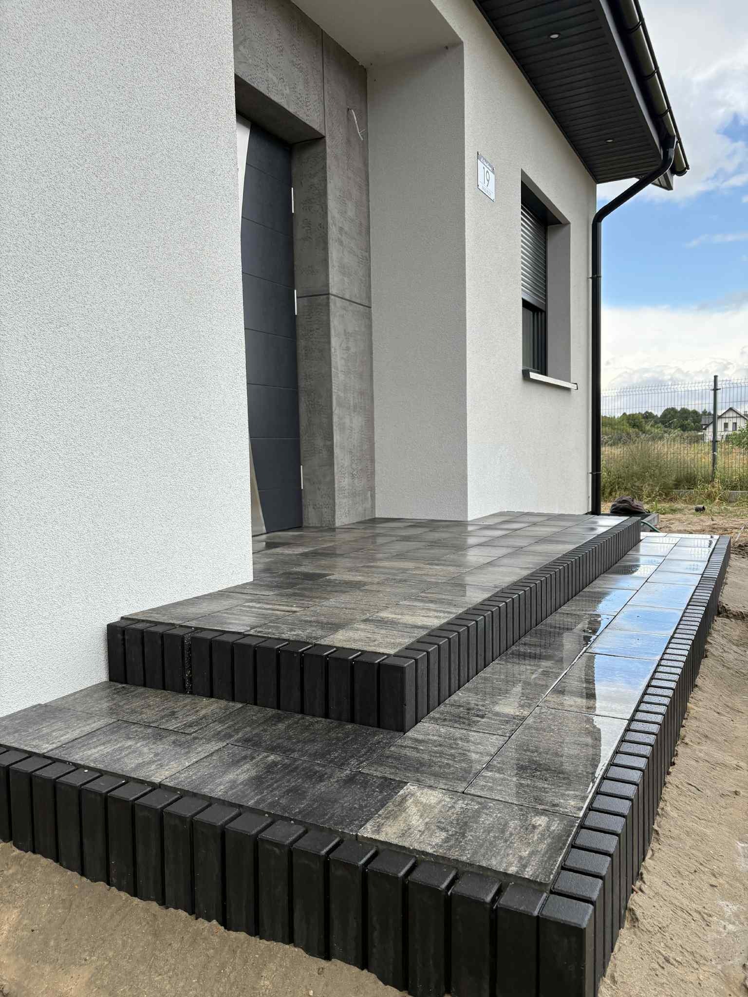 Nowoczesne schody wejściowe z kostki brukowej w odcieniach szarości, wykończone czarnym obrzeżem. Widoczny fragment elewacji domu z numerem 19 i szare drzwi.