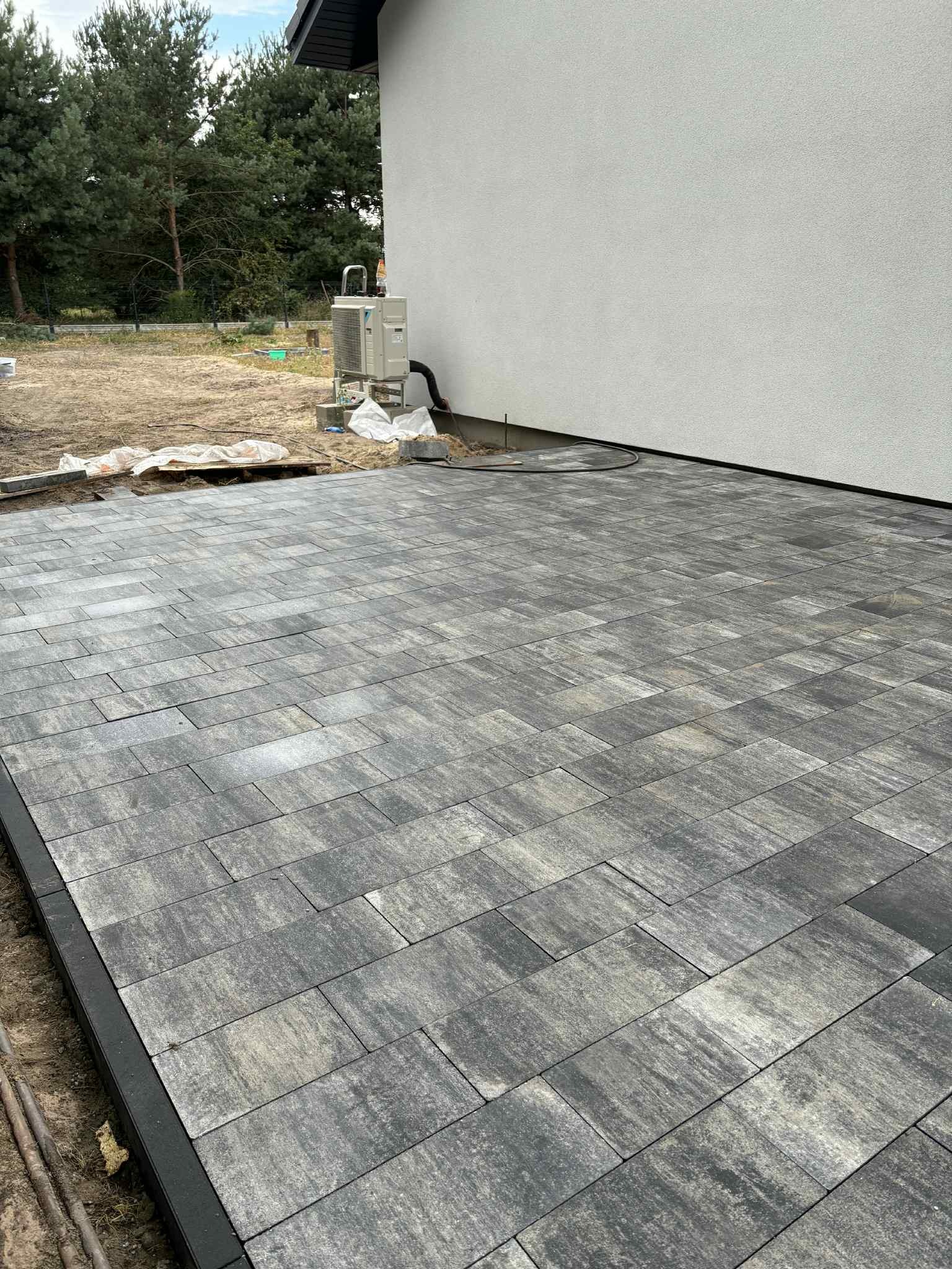 Ułożona, szara kostka brukowa z czarnym obrzeżem przy ścianie budynku z klimatyzatorem. Częściowo przygotowany teren pod dalsze prace ziemne.