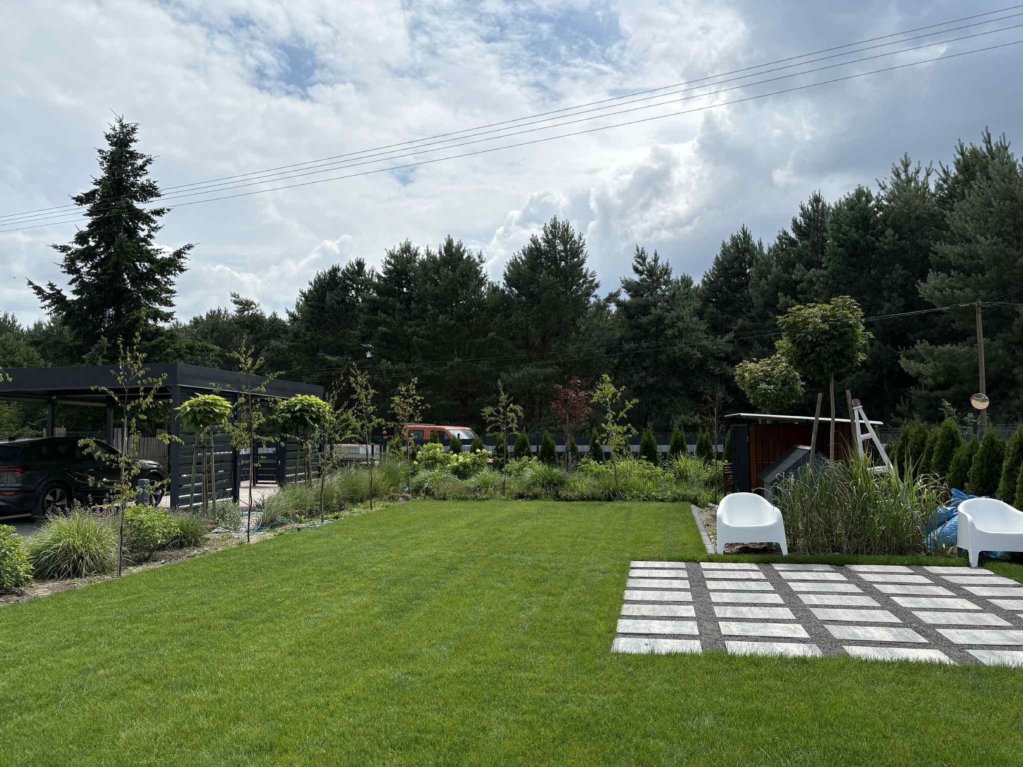 Zadbany ogród z równo przystrzyżonym trawnikiem, dekoracyjnymi drzewami, kostką i białymi krzesłami na tle zieleni i pochmurnego nieba. W tle zaparkowany czarny samochód.