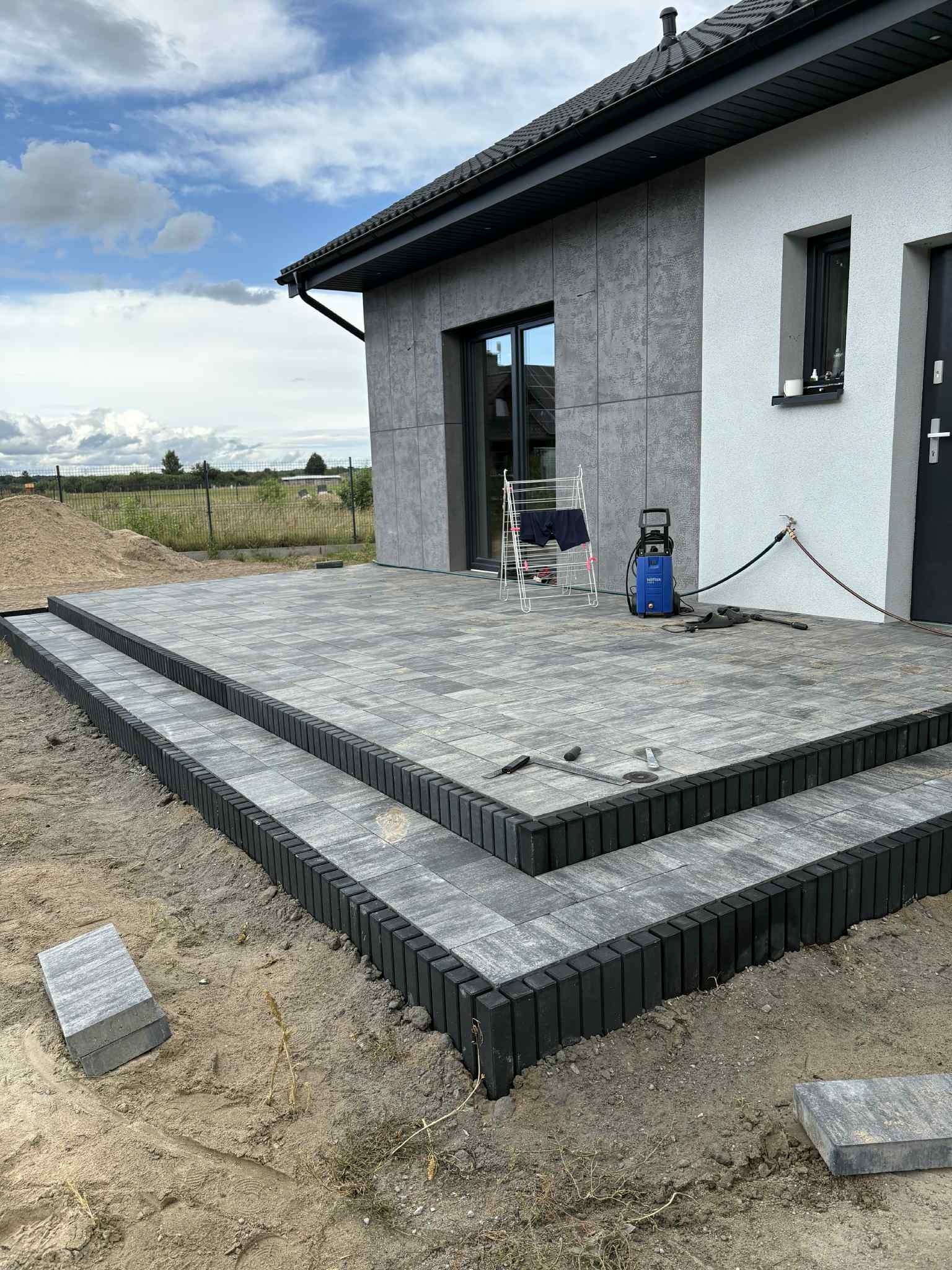 Ułożony taras z kostki brukowej w odcieniach szarości, z dwoma stopniami obrzeżowanymi czarnym krawężnikiem, na tle nowoczesnego domu i błękitnego nieba.