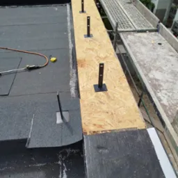 Montaż płaskiego dachu z papy termozgrzewalnej, widoczne mocowania do instalacji odgromowej i narzędzia na powierzchni, rusztowanie w tle.