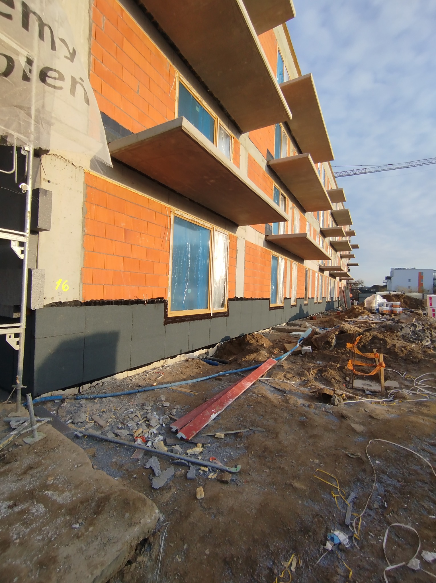 Budynek w trakcie budowy: widoczne cegły, okna zabezpieczone folią, izolacja termiczna przy gruncie, rusztowania balkonowe, plac budowy z gruzem i materiałami budowlanymi.