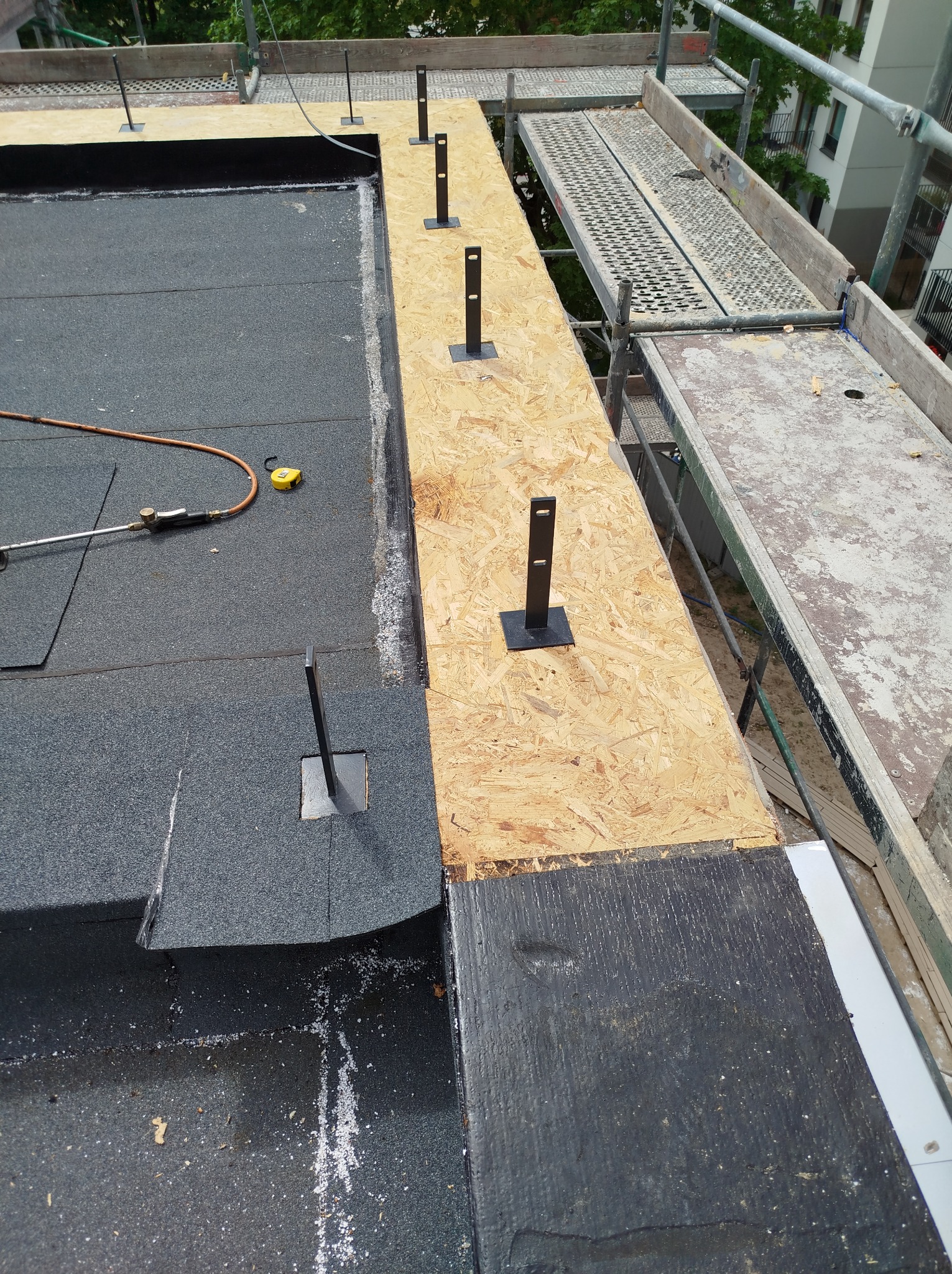 Montaż płaskiego dachu z papy termozgrzewalnej, widoczne mocowania do instalacji odgromowej i narzędzia na powierzchni, rusztowanie w tle.