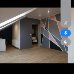 Pokój na poddaszu po remoncie: drewniana podłoga, ściana z geometrycznym wzorem w odcieniach szarości, różu i czerni, widoczne wejście do łazienki z umywalką.