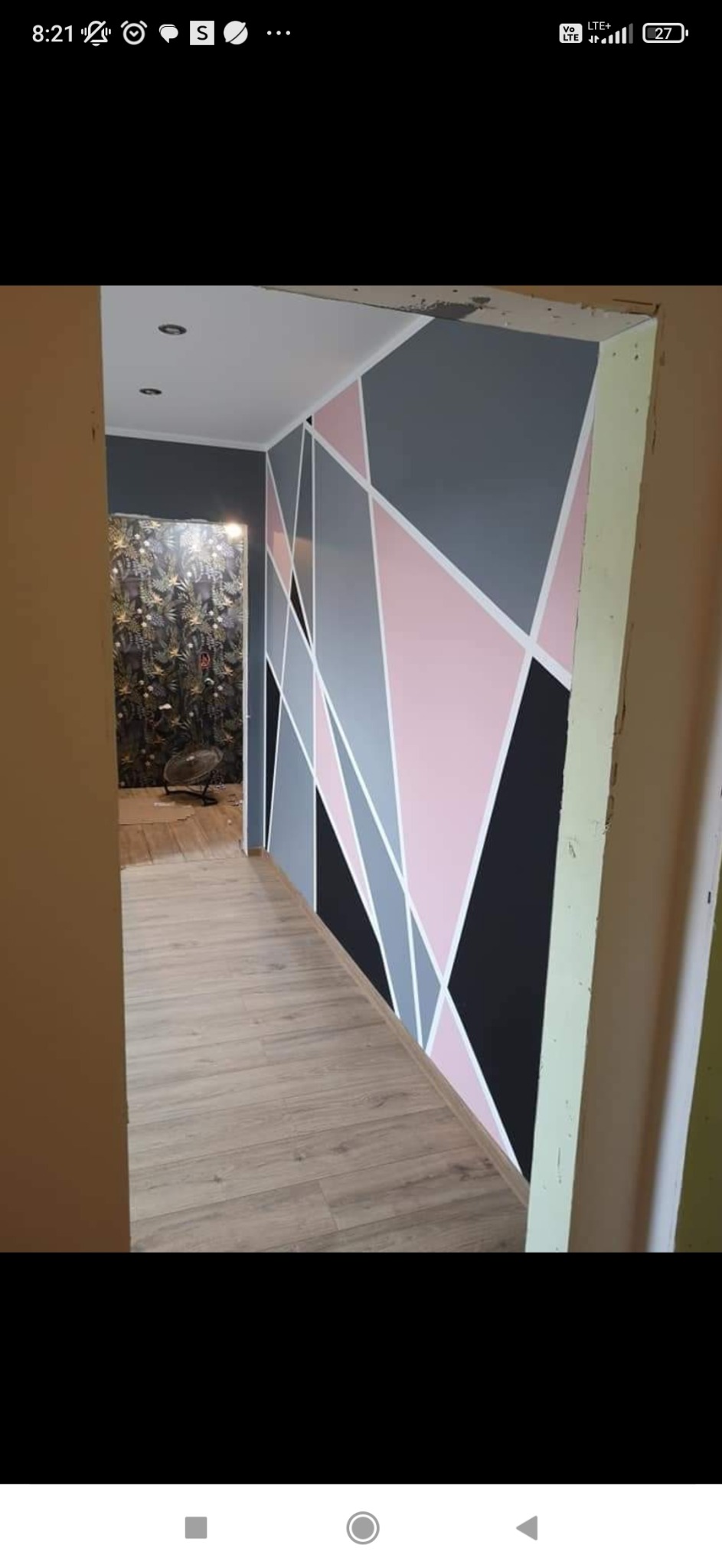 Ściana w korytarzu z geometrycznym wzorem w odcieniach szarości, różu i czerni, widoczne panele podłogowe i fragment tapety na ścianie w głębi.