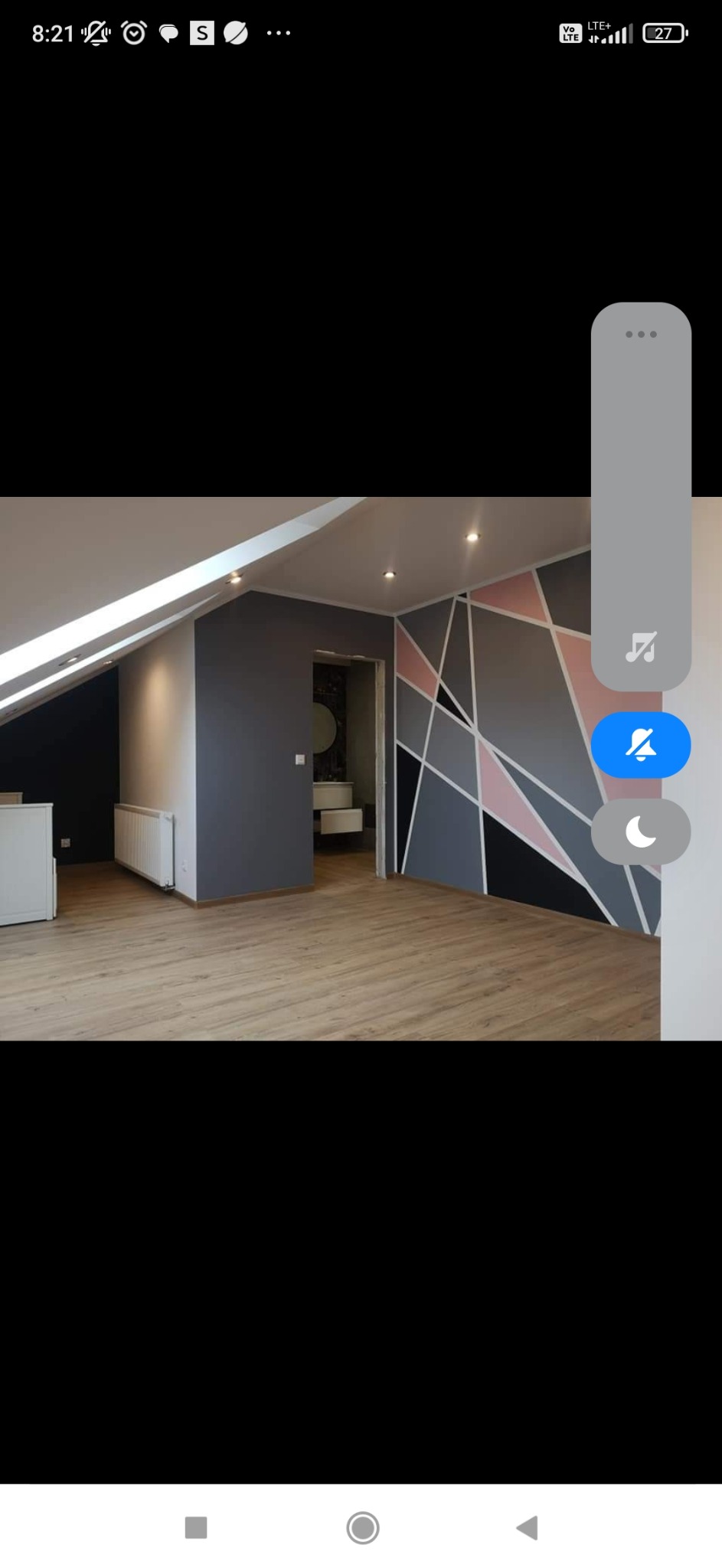 Pokój na poddaszu po remoncie: drewniana podłoga, ściana z geometrycznym wzorem w odcieniach szarości, różu i czerni, widoczne wejście do łazienki z umywalką.