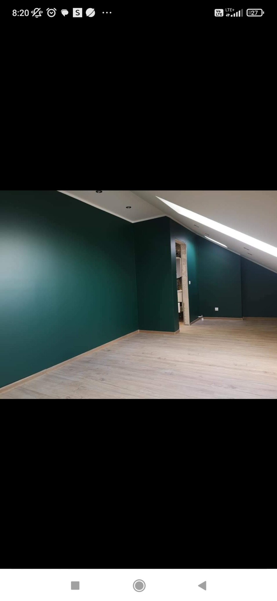 Pomalowane na ciemnozielono ściany w pokoju na poddaszu z jasnymi panelami podłogowymi i widocznym fragmentem ościeżnicy drzwiowej.