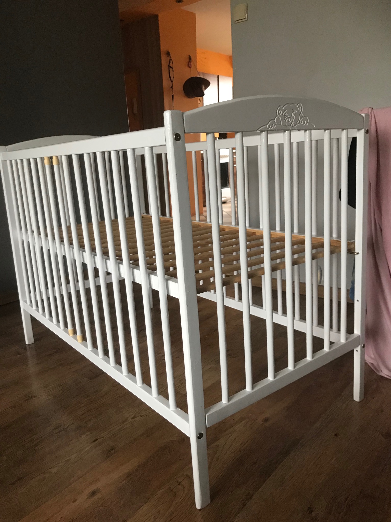 Białe łóżeczko dziecięce z drewnianym stelażem, widoczne z boku na tle drewnianej podłogi i fragmentu pokoju.