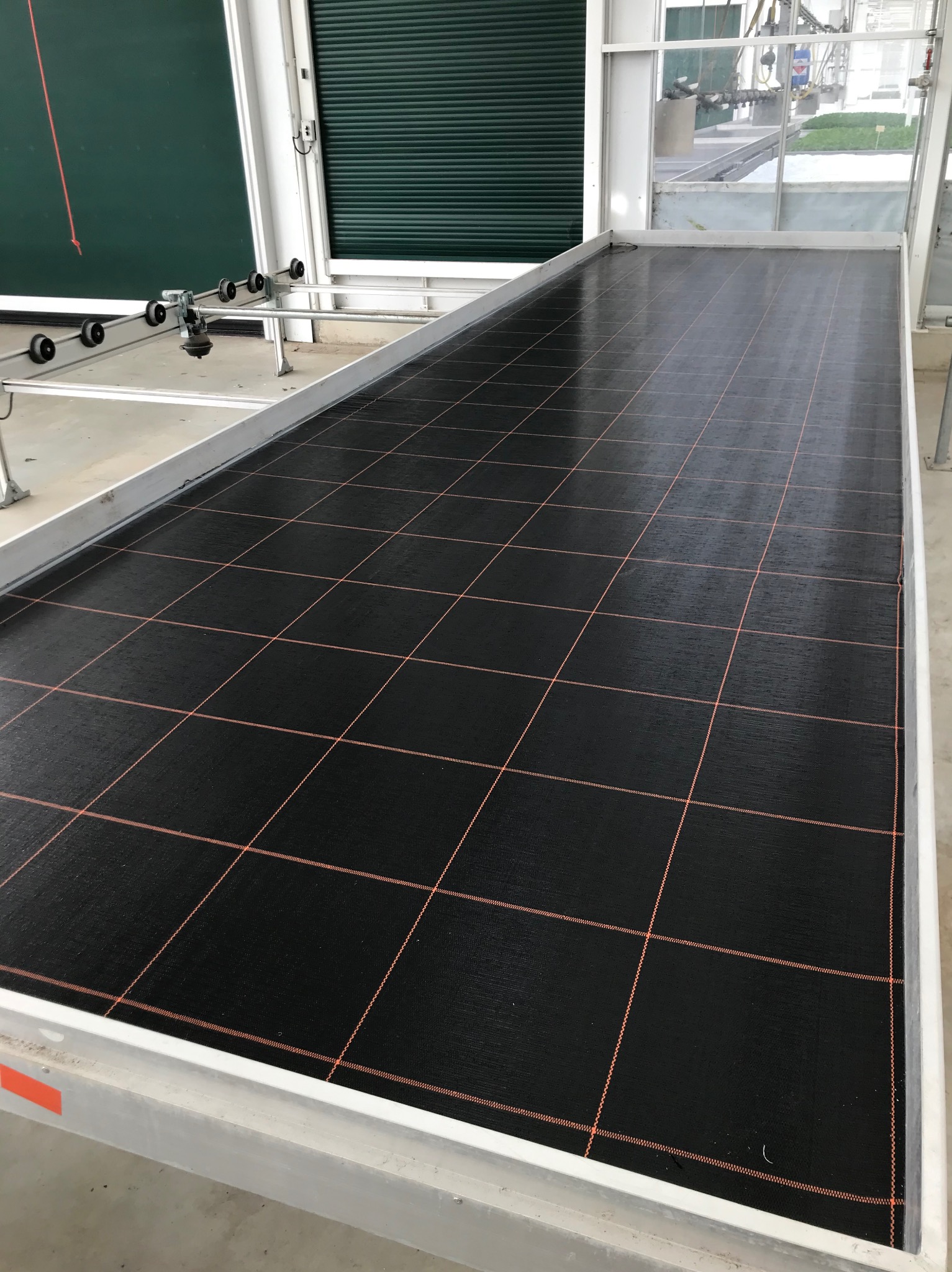 Metalowy stół z czarną, siatkową powierzchnią w szklarni, widoczne rolki i system nawadniania w tle.