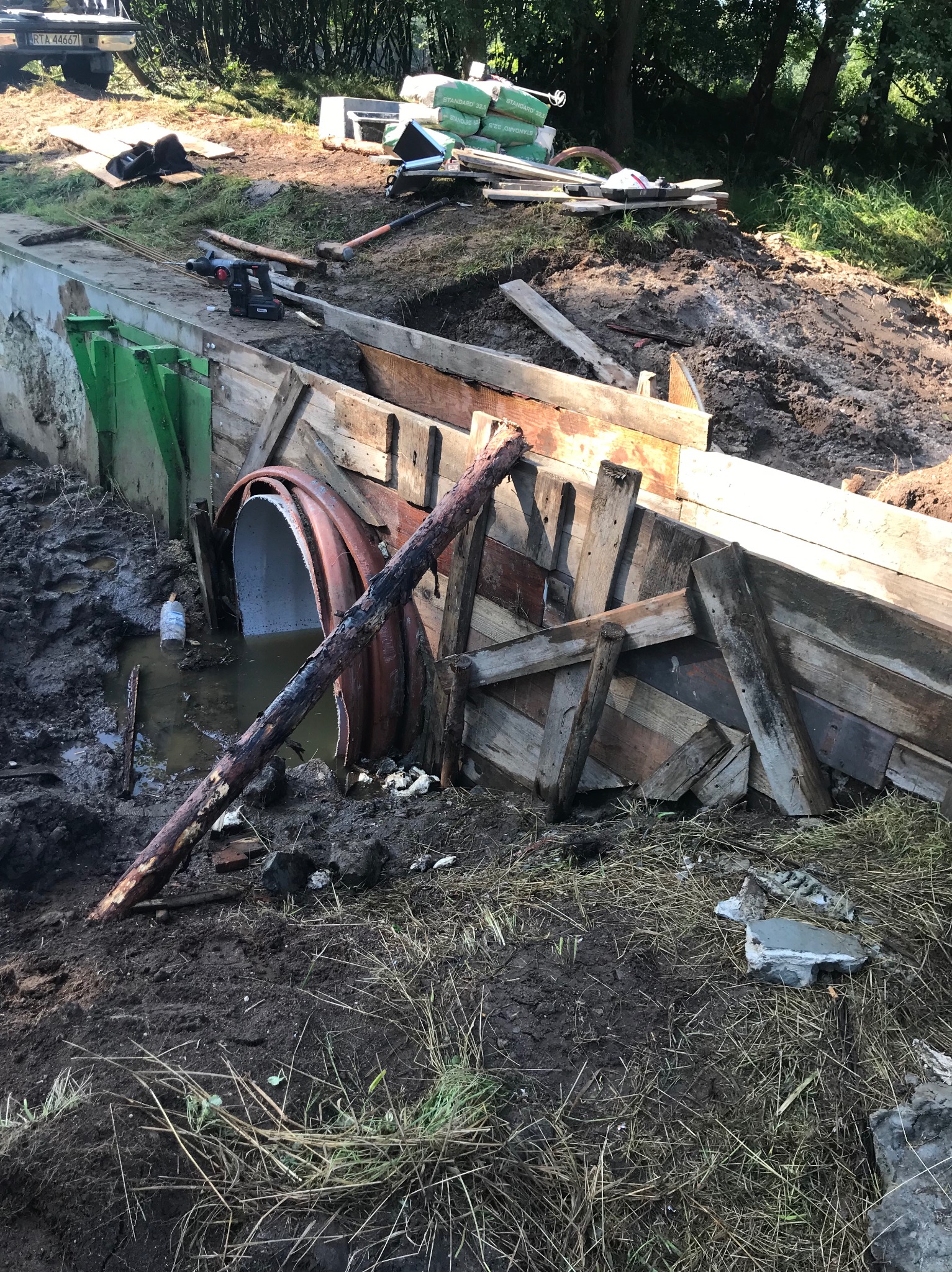 Budowa przepustu: rura w ziemi otoczona drewnianą konstrukcją, narzędzia i materiały budowlane w tle, widoczne ślady prac ziemnych.