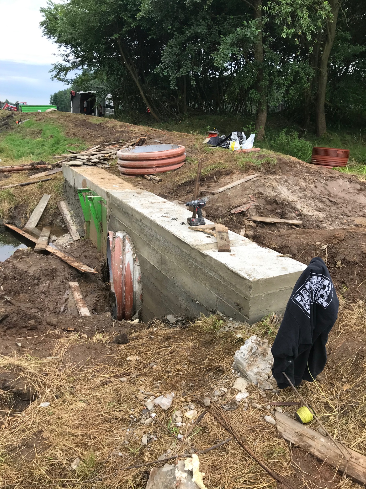 Betonowy przepust z zielonymi elementami konstrukcyjnymi w trakcie budowy, widoczna woda, narzędzia i elementy budowlane w otoczeniu.