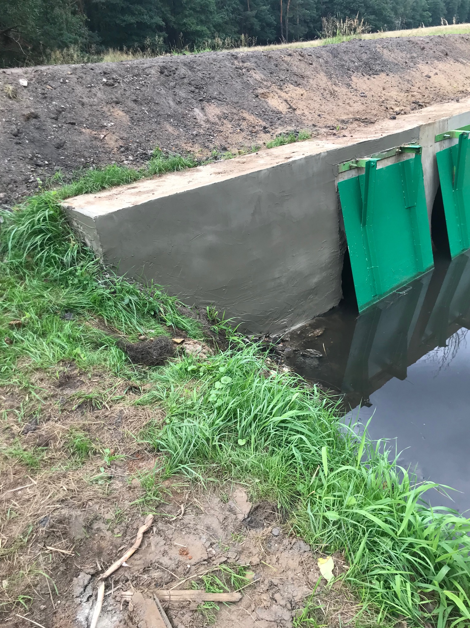 Świeżo wyremontowana betonowa konstrukcja przepustu wodnego z zielonymi metalowymi zasuwami, otoczona ziemią i bujną trawą.