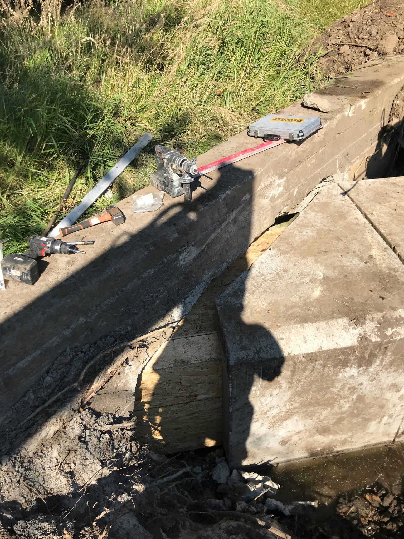 Budowa betonowego obrzeża, widoczne narzędzia: wiertarka, młotek, poziomica laserowa w metalowej walizce, ziemia i trawa w tle.