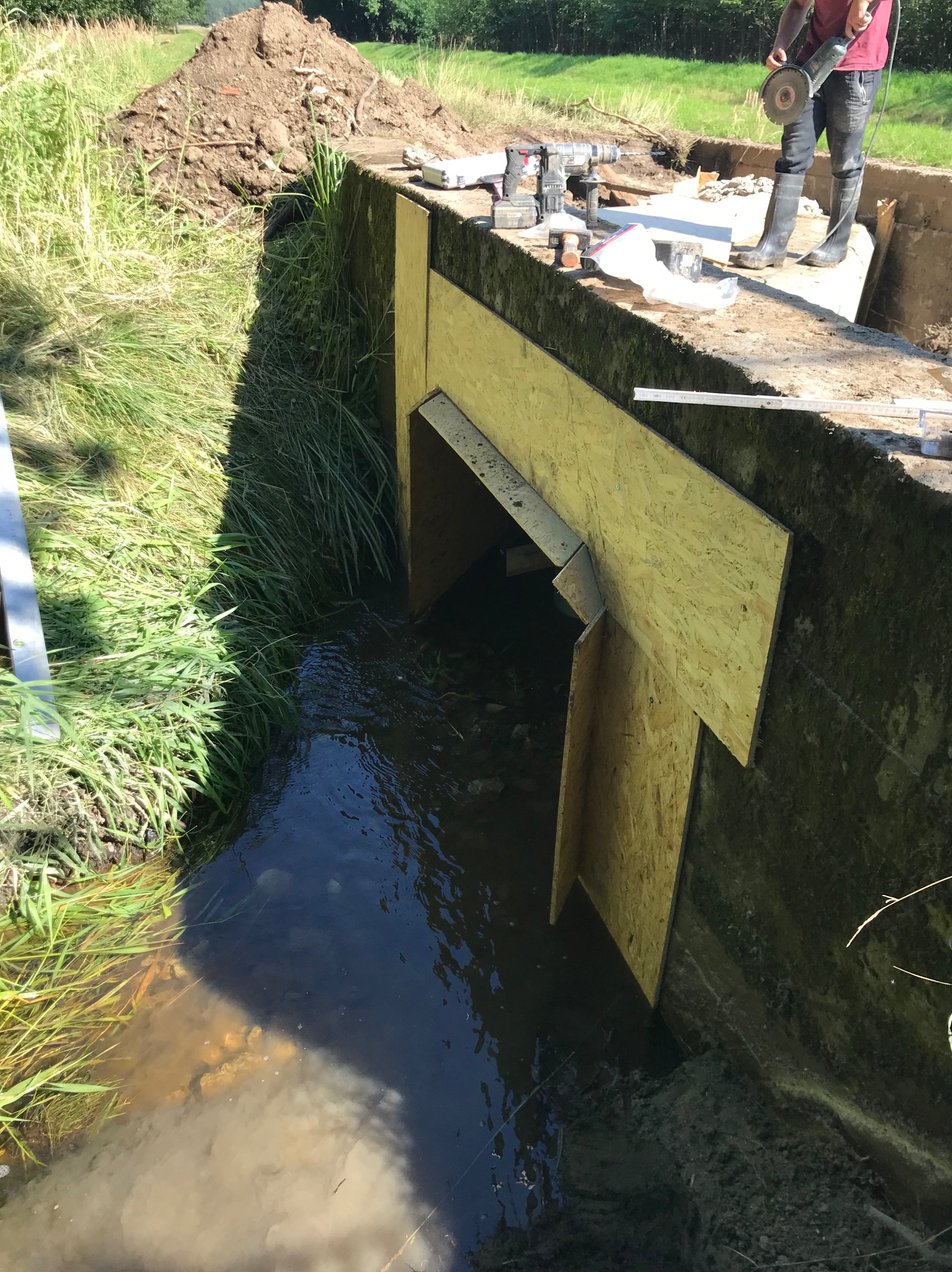 Budowa przepustu wodnego z drewnianą szalunką, widoczne narzędzia i pracownik w gumowych butach podczas cięcia metalu szlifierką kątową.