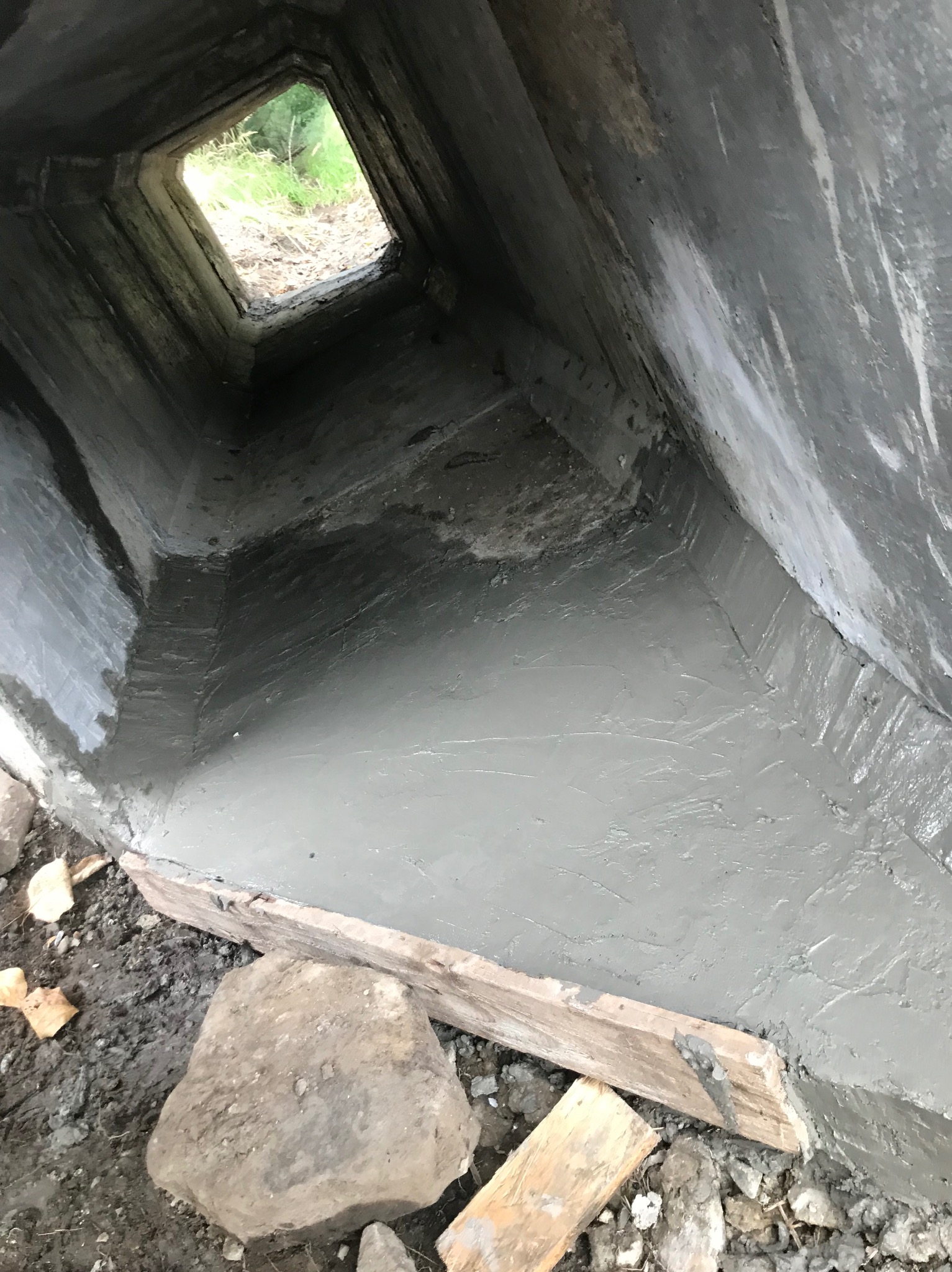 Wnętrze betonowego przepustu po wylaniu świeżej warstwy betonu, widoczne drewniane szalunki i kamienie na zewnątrz, w oddali otwór wylotowy przepustu z zielenią.