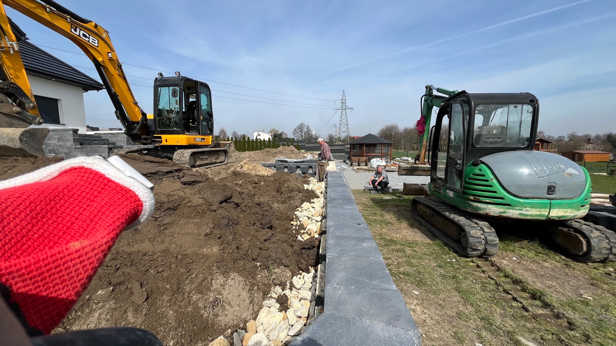 Dwie koparki, żółta i zielona, pracują nad wyrównywaniem terenu i układaniem kamiennych murków w Pisarzowicach, widok z perspektywy pracownika w czerwonej rękawicy.