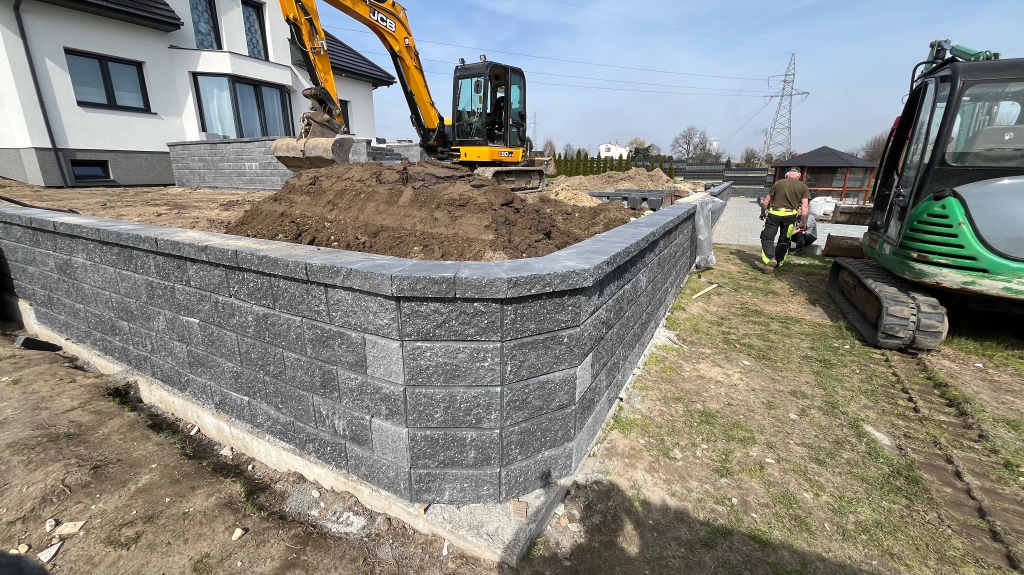 Budowa muru oporowego z szarych betonowych bloczków; w tle żółta koparka i zielona mini koparka oraz pracownicy.