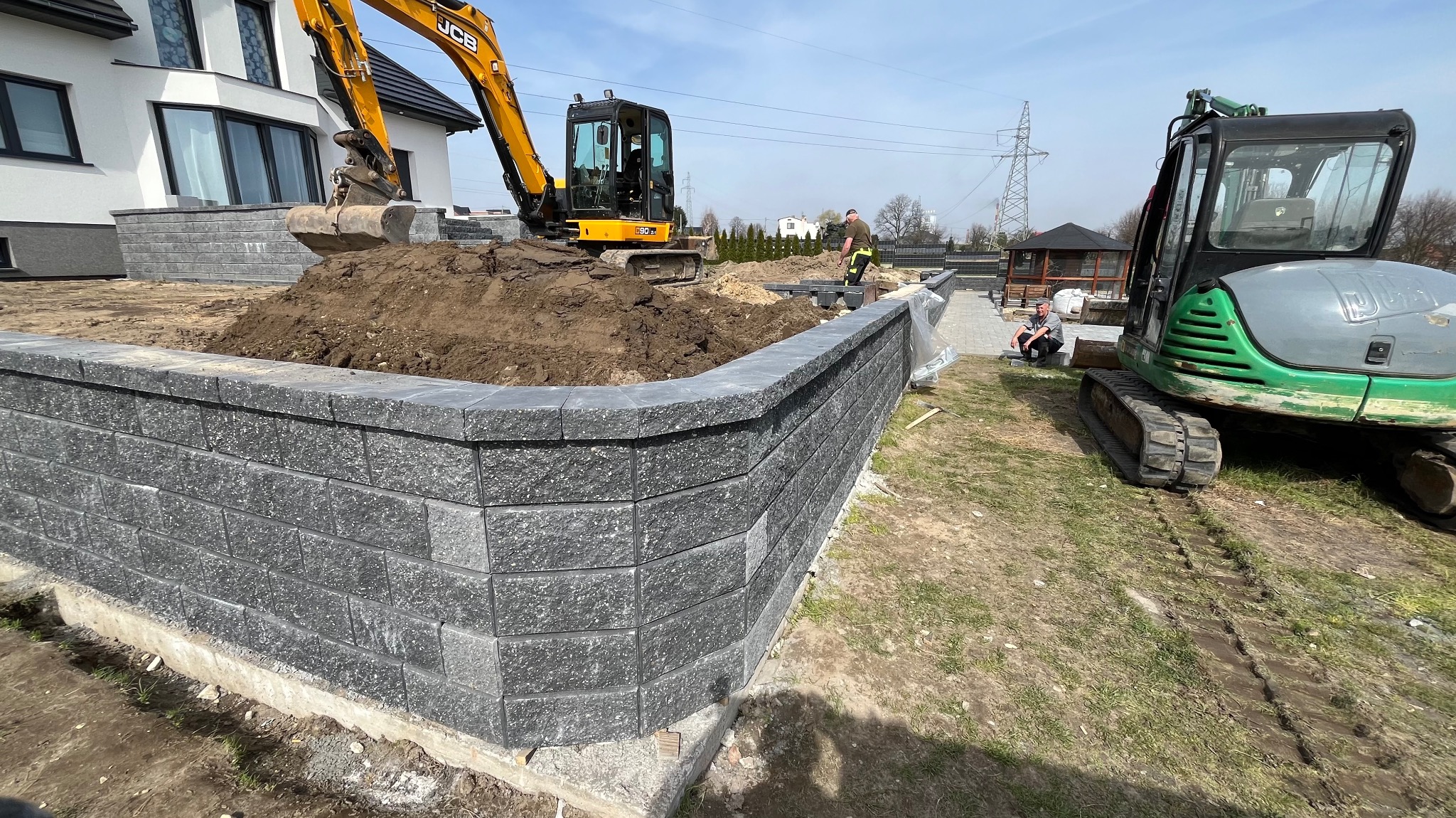 Budowa muru oporowego z szarej kostki betonowej na terenie posesji, z użyciem koparki JCB i mniejszej koparki, widoczni pracownicy w trakcie robót ziemnych i układania kostki.