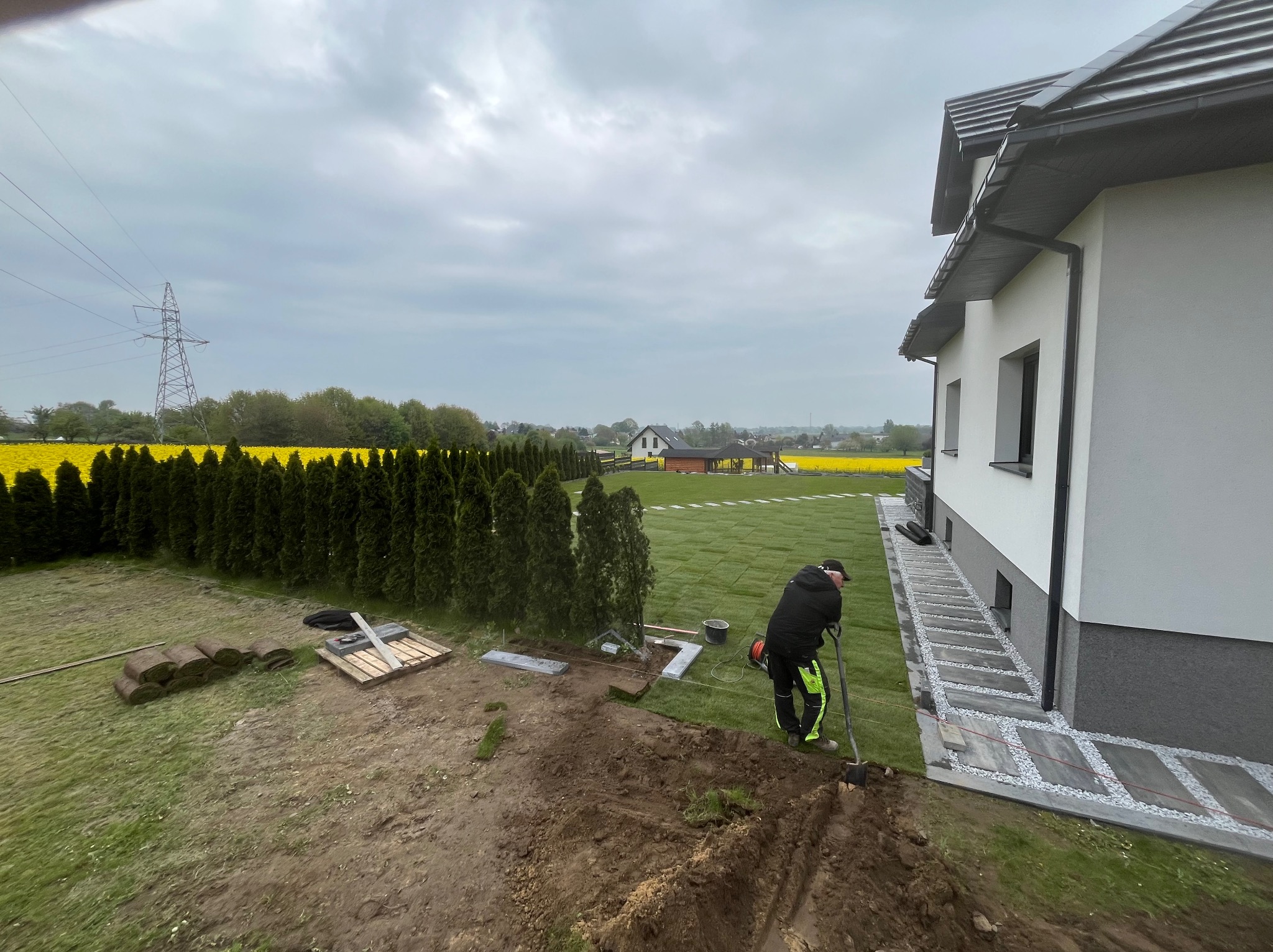 Układanie trawnika z rolki przy nowoczesnym domu; widoczne rolki trawy, przygotowany teren, pracownik z łopatą i ułożona ścieżka z płyt.
