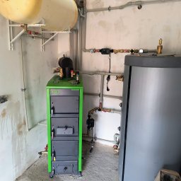 PIPEPOWER Marcin Linowski - Zielony piec SAS, szary zbiornik, biały zasobnik i rury w kotłowni w trakcie montażu instalacji grzewczej. Widoczne ślady na ścianie po pracach instalacyjnych.