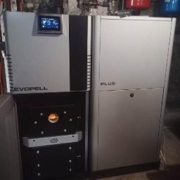 PIPEPOWER Marcin Linowski - Nowoczesny, srebrny piec Evopell Plus z otwartymi drzwiczkami paleniska, widoczne rury instalacji grzewczej i zbiorniki wyrównawcze w tle.