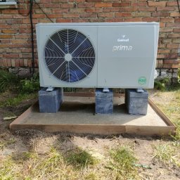 PIPEPOWER Marcin Linowski - Biała pompa ciepła Galmet Prima R32, ustawiona na betonowych bloczkach, na przygotowanym podłożu z piasku i drewnianej ramy, przy ceglanej ścianie budynku.