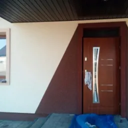 Elewacja domu z oknem i drzwiami wejściowymi, wykończona w dwóch odcieniach beżu i brązu. Widoczny fragment podbitki dachowej i schody wejściowe.