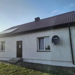 Dom jednorodzinny z panelami słonecznymi na dachu i anteną satelitarną na elewacji. Widoczne okna z białymi ramami i brązowe rynny.