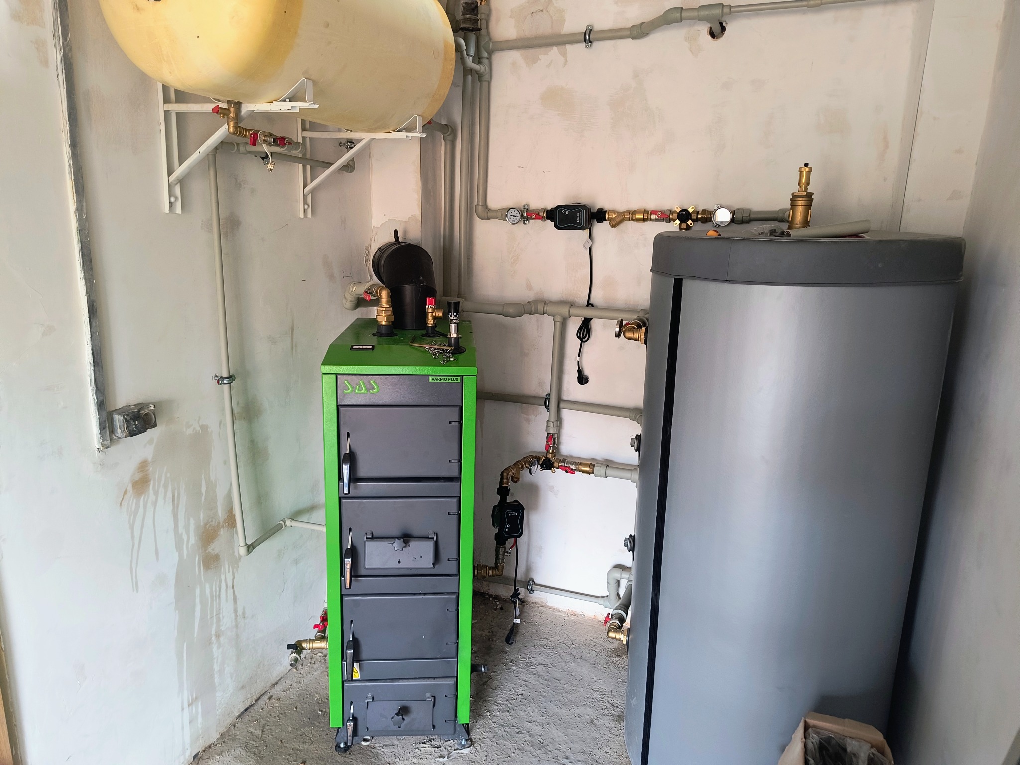 Zielony piec SAS, szary zbiornik, biały zasobnik i rury w kotłowni w trakcie montażu instalacji grzewczej. Widoczne ślady na ścianie po pracach instalacyjnych.