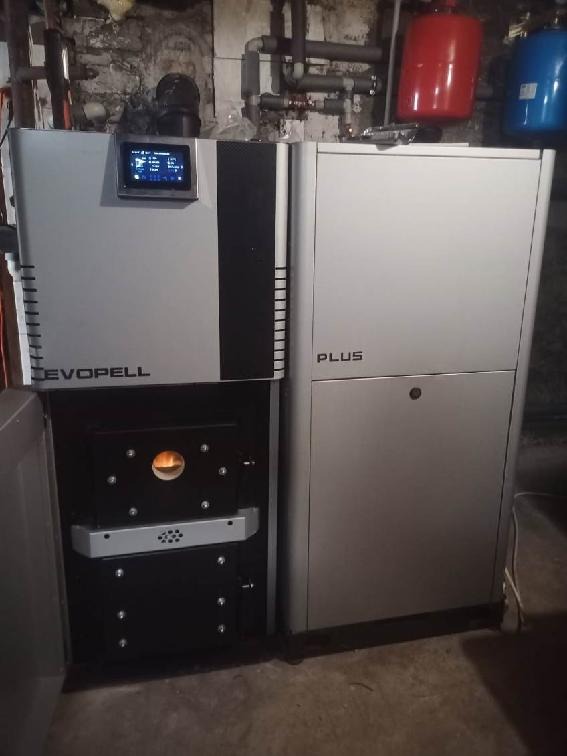 Nowoczesny, srebrny piec Evopell Plus z otwartymi drzwiczkami paleniska, widoczne rury instalacji grzewczej i zbiorniki wyrównawcze w tle.