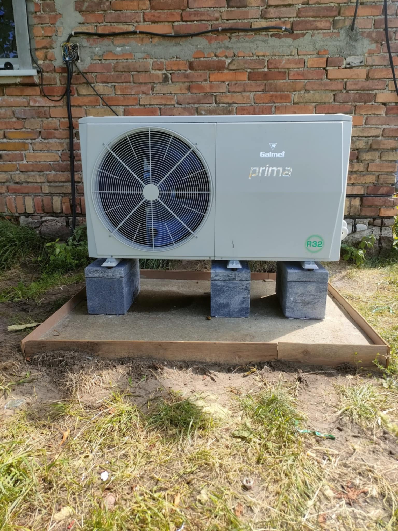 Biała pompa ciepła Galmet Prima R32, ustawiona na betonowych bloczkach, na przygotowanym podłożu z piasku i drewnianej ramy, przy ceglanej ścianie budynku.