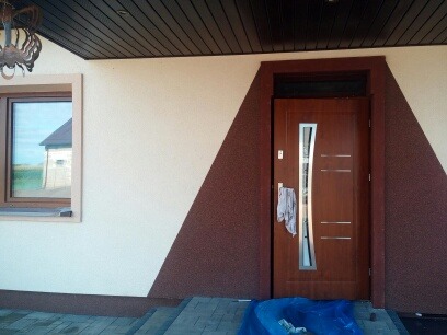 Elewacja domu z oknem i drzwiami wejściowymi, wykończona w dwóch odcieniach beżu i brązu. Widoczny fragment podbitki dachowej i schody wejściowe.