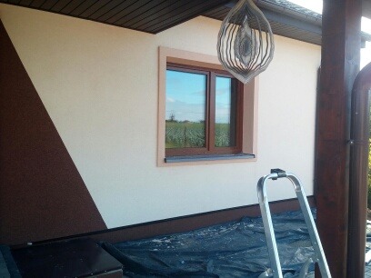 Elewacja domu po malowaniu z dekoracyjnym brązowym pasem. Widoczne okno z brązową ramą i drabina oparta o ścianę. Całość w jasnych, naturalnych barwach.