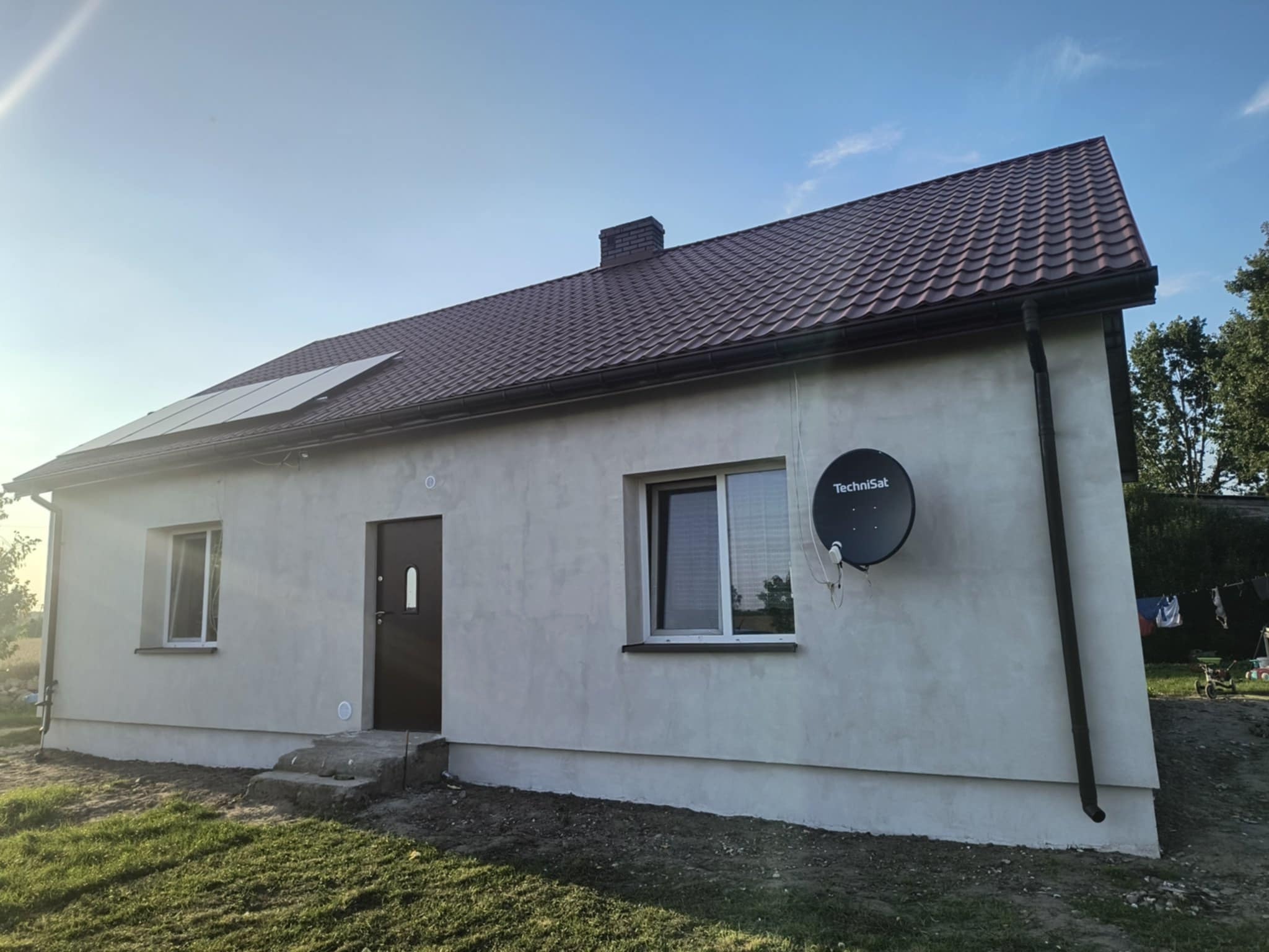 Dom jednorodzinny z panelami słonecznymi na dachu i anteną satelitarną na elewacji. Widoczne okna z białymi ramami i brązowe rynny.