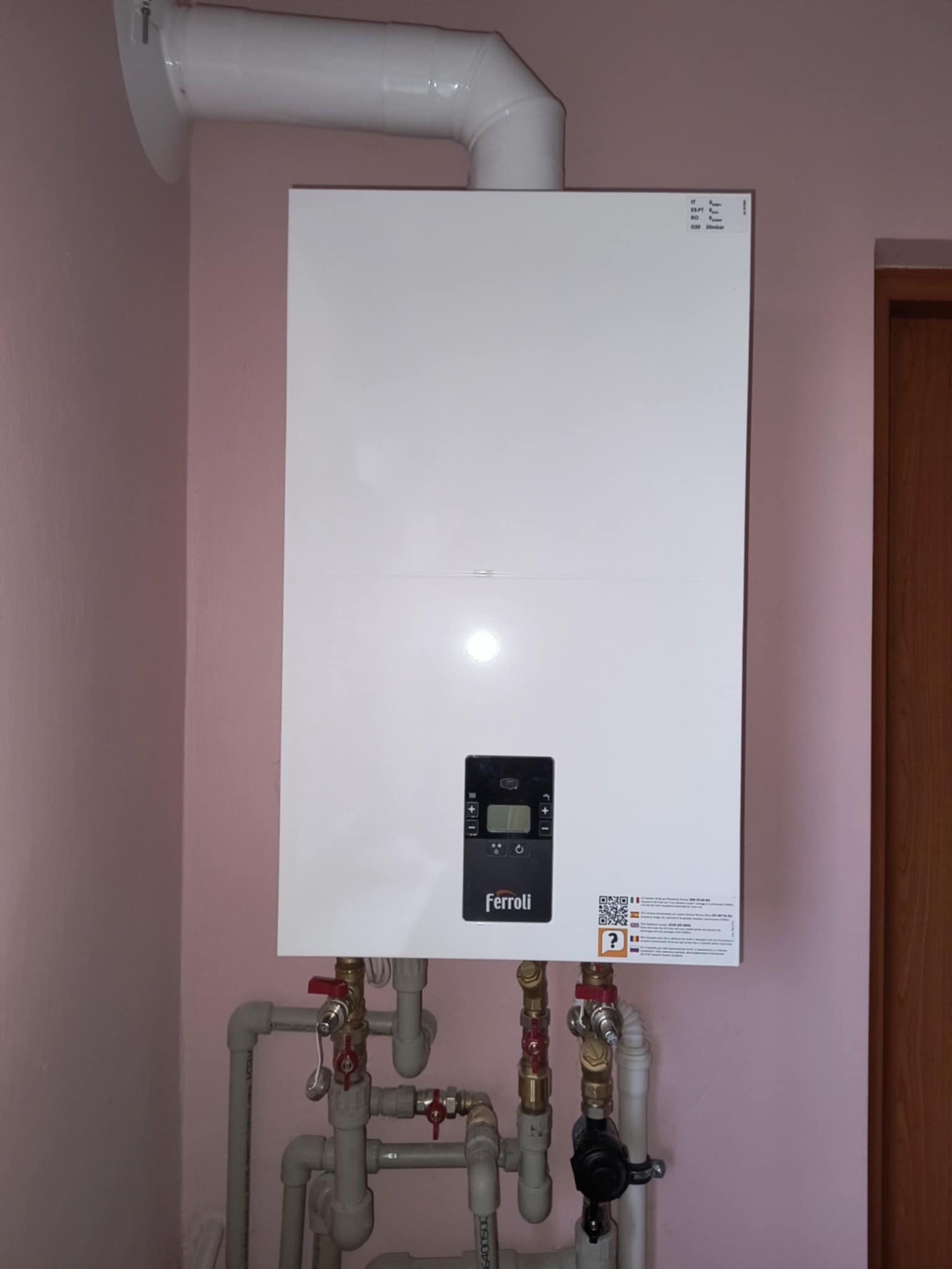 Biały, wiszący piec gazowy Ferroli z szarymi rurami i zaworami na tle różowej ściany. Widoczny komin odprowadzający spaliny.