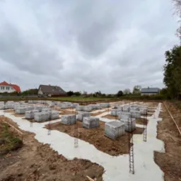 Przygotowany teren pod budowę domu z wylanymi fundamentami i zbrojeniami, na których ułożone są bloczki fundamentowe Durabet w opakowaniach foliowych, widoczne drewniane szalunki, w tle zabudowa...