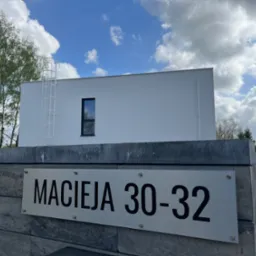 Nowoczesny, biały budynek z płaskim dachem i wąskim oknem, widoczny zza szarego muru z tabliczką adresową 'Macieja 30-32', na tle błękitnego nieba z chmurami.